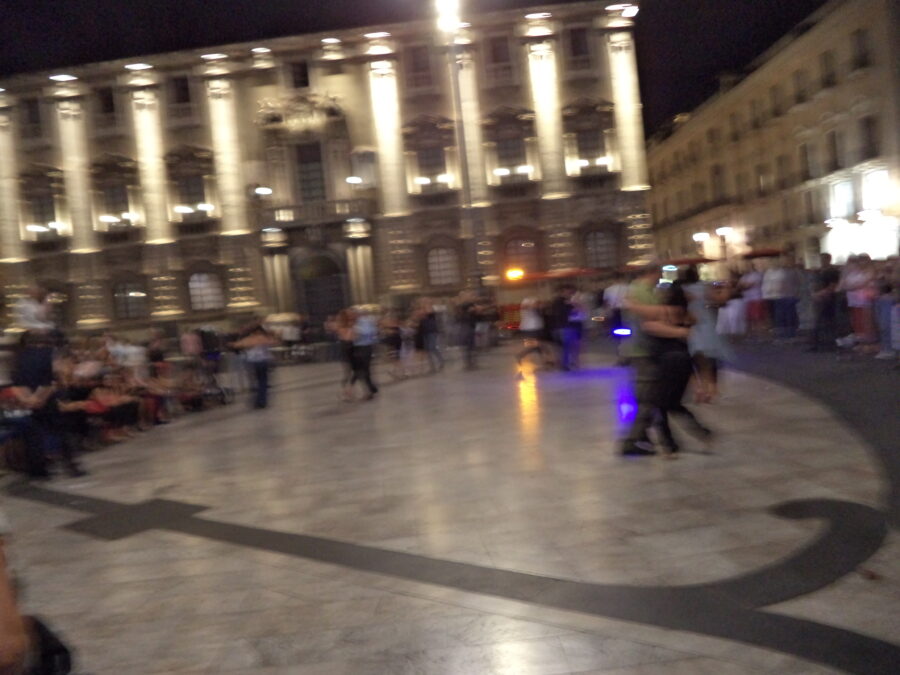 A slightly blurry view of Piazza dell Duomo in the evening. In the centre of the shot people are dancing in pairs. Around the edges people are standing and sitting watching the dancers. Historic buildings are lit-up in the background.