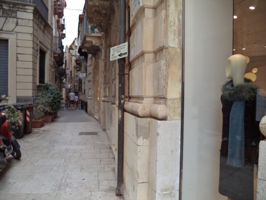 A narrow side-street on Ortigia, the historic centre of Syracuse. A mannikin can be seen in a clothes shop window in the foreground. Many of the buildings house shops and small businesses at street level. There are narrow stone balconies on the upper storeys.
