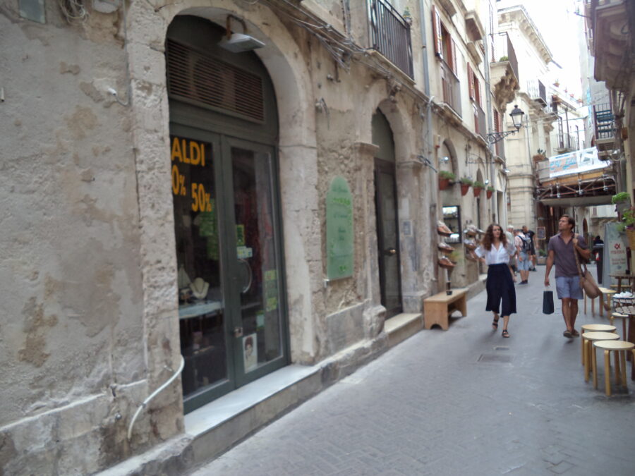 A narrow side-street on Ortigia, the historic centre of Syracuse. A mannikin can be seen in a clothes shop window in the foreground. Many of the buildings house shops and small businesses at street level. There are narrow stone balconies on the upper storeys.