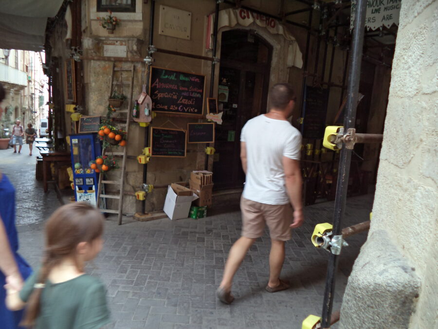 At the junction of two narrow streets in Ortigia. A family are passing by in front. To the right the street is covered by scaffolding. There is a cafe or bar in the building ahead. There are chalkboards advertising offers or specialities. An old wooden ladder is decoratively placed against a wall with baskets attached.