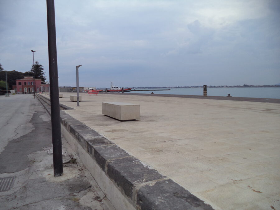 Sea front promenade on Ortigia. The wide promenade is paved with slabs. At the far end is a red painted building. A pair of small boats are moored nearby. The sea appears calm. The coast south of Syracuse can be seen spread-out in the distance.