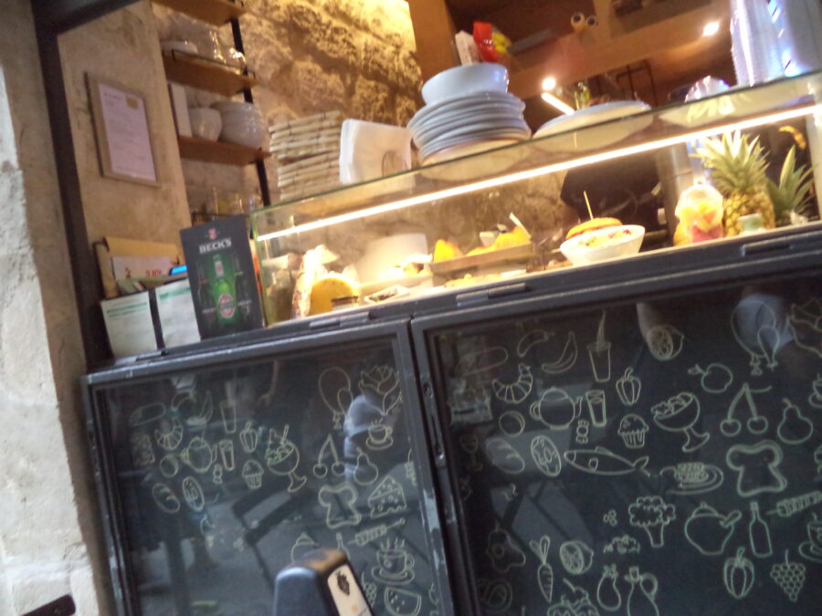 A street-side counter belonging to a café or restaurant. Stacks of plates and table mats sit on top. Food is visible through the glass front of the counter including a burger and a pineapple. The lower part of the counter has black panels decorated with line-drawn food illustrations like cherries, fish and cupcakes.