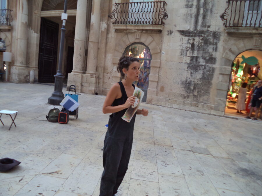 A young woman standing in Piazza Duomo. She is holding a drawing and appears to be some sort of street artist or performer. There is a folding stool and a black hat on the ground behind her. An historic stone building can be seen in the background.