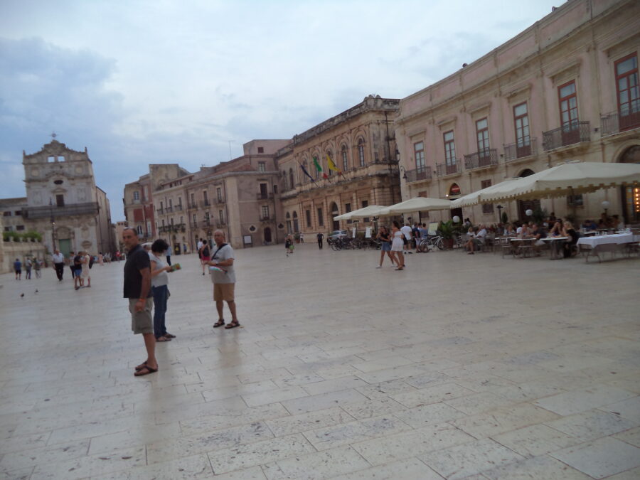 Another view across Piazza Duomo. The Church of Santa Lucia alla Badia is again visible ahead. There are outdoor restaurant tables shaded by including grand residences or palazzi surround the square.