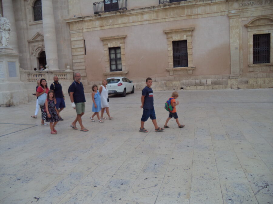 Looking towards the east side of Piazza Duomo. A small part of Syracuse Cathedral can be seen to the left including a statue of St Peter standing on a plinth at the end of a stone balustrade. Corinthian columns can also be seen. A group of adults and children are passing by in front.