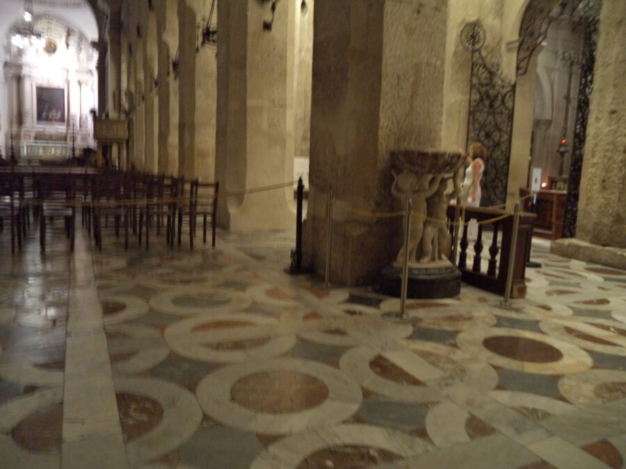 Inside Syracuse Cathedral looking down the nave with the altar vaguely visible ahead. A row of substantial stone columns supporting Norman arches can be seen to the right of the nave. Further to the right side, a large elaborate wrought iron door leading into a side chapel can be seen. In front of the nearest large column is a stone font: two small children are sculpted holding up a large bowl. The marble floor forms a geometric pattern of circles and straight lines. There are rows of wooden chairs.