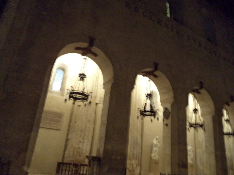 Looking up from the nave of Syracuse Cathedral. A row of tall Norman arches can be seen. Within the centre of each arch wrought-iron chandeliers hang. Statues can vaguely be seen along the adjoining side aisle.