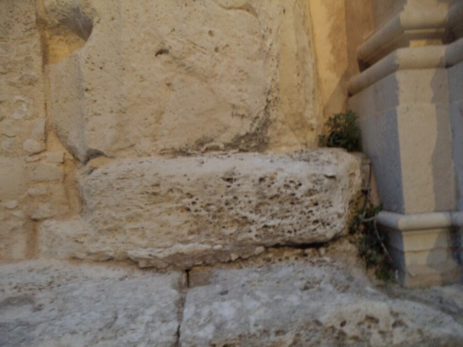 Large weathered pale stone blocks at the base of Syracuse Cathedral's outside walls. A few small green plants are growing in a shaded corner. These blocks may originate from the Greek temple that once stood on this site.