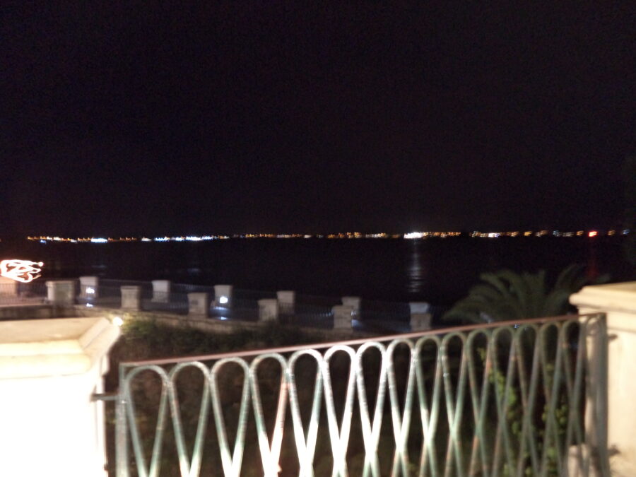 Night time sea view from Ortigia. A line of distant lights on the opposite side of the bay illuminate the horizon and reflect on the water which appears almost black in darkness. There is a terrace in the foreground with walls and railings.
