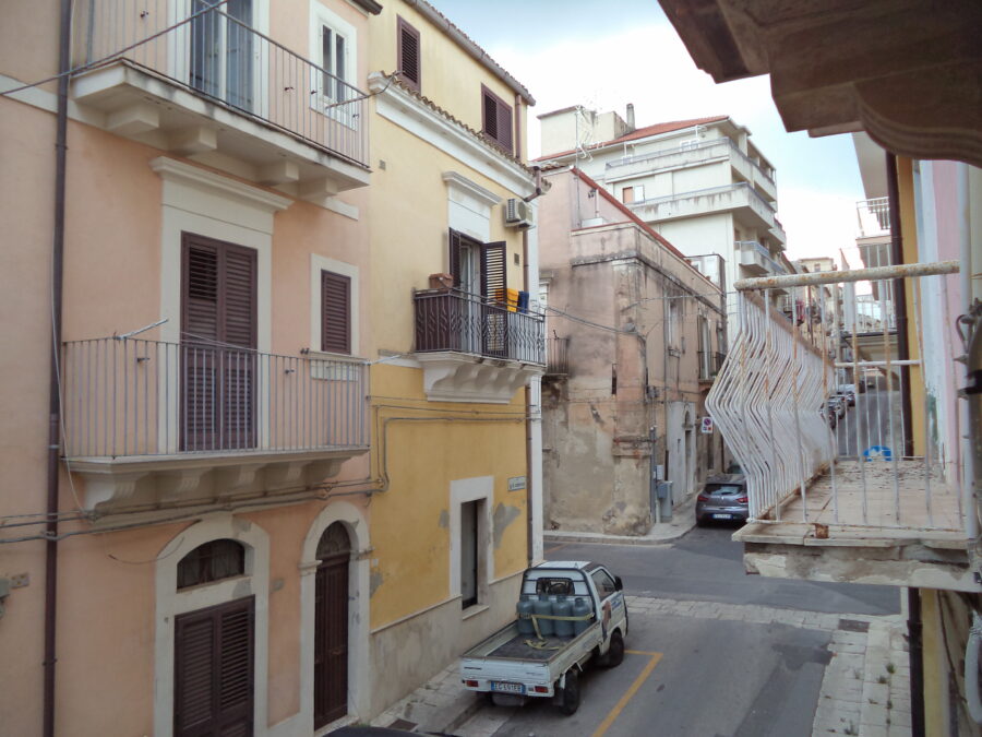 Looking the other way, uphill along the residential street from the hotel balcony. The balconies belonging to adjoining houses and apartments can be seen. The houses opposite  are painted in pastel colours in yellow and pink. 