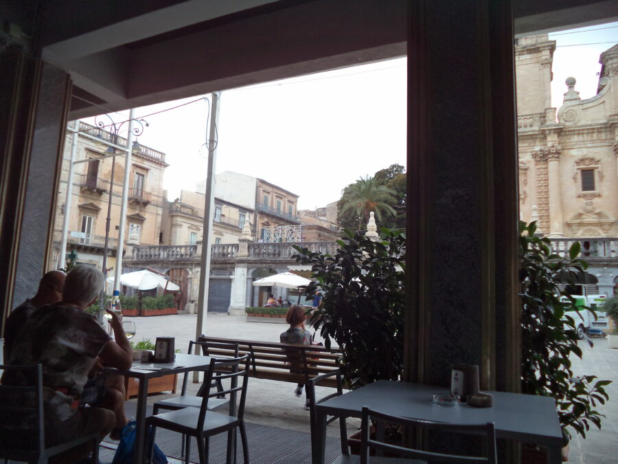 View from a cafe overlooking Piazza San Giovanni, a square sited in front of Ragusa Cathedral. The front of the cathedral is raised up on a terrace above and only a small part of the front façade is visible. People can be seen sitting on benches or at cafe tables in the square.