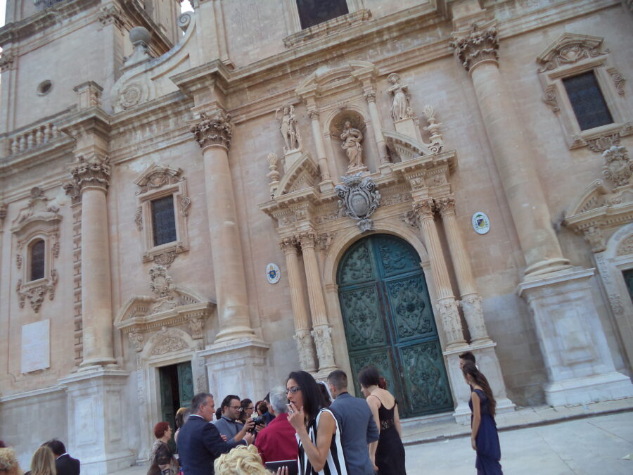 The Baroque front façade of Ragusa Cathedral (also known as the Cathedral of San Giovanni Battista). The cathedral is dedicated to Saint John the Baptist and was constructed after the 1693 Sicily earthquake. An earlier church dedicated to John the Baptist at a different location was destroyed by the earthquake. The church became a cathedral in 1950. The photo shows the lower part of the façade including the large main doorway. The cathedral is elaborately decorated with Corinthian columns, statues and floral decoration around the windows. The arched main doorway has two Corinthian columns at each side and a carved motif or crest above with cherubs at the sides and a crown on top. Above this there is more carved stonework including three statues. In the foreground, a group of people are assembled, they are part of a wedding party.
