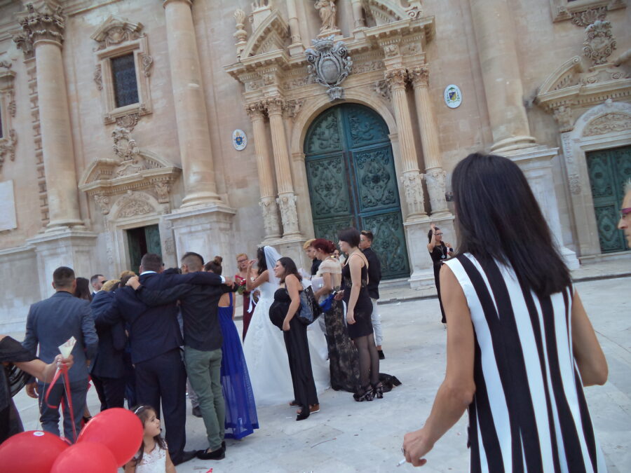 Another shot of Ragusa Cathedral with a wedding group assembled in the square in front. The bride can be seen dressed in a white gown with a veil. Other people are dressed formally in suits and dresses. Someone is holding red balloons in the foreground.