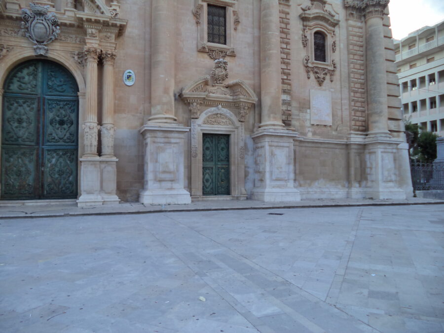 The Baroque front façade of Ragusa Cathedral. The shot is looking towards the right side, showing the main doorway, and another smaller panels containing a curved geometric design. The doors are painted turquoise-green. The massive bases belonging to Corinthian columns jut out at ground level.