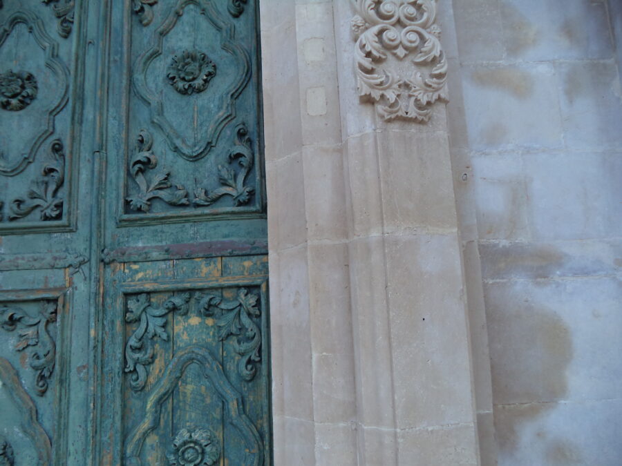 Another view of Ragusa Cathedral's side door. The door has four panels with a flower carved in the centre of each and leaf designs in the corners. Some of the carved stonework around the door can also be seen. This is again a curvy design resembling leaves.