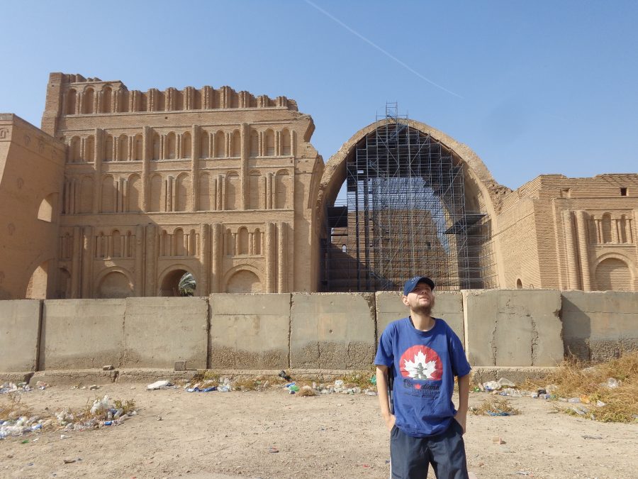 View of Taq Kasra with the huge ancient brick arch towards the centre and tall brick walls at either side that are adorned with rows of arched alcoves. The central arch extends back to form a hall. It is more than 30 metres (110 ft) in height. Originally it covered an area of 24 metres (80 ft) in width by 48 metres (160 ft) in length and formed part of an imperial palace complex. The throne room is thought to have been under or behind the arch. The photo shows the interior of the arch filled with scaffolding, perhaps for restoration work, or simply to reduce the risk of it collapsing. The surrounding walls rise up to 37 metres (121 feet) in height. Tony is stood in the foreground with his hands in his pockets. Immediately behind him is a concrete barrier and the surrounding area is scattered with litter and weeds.