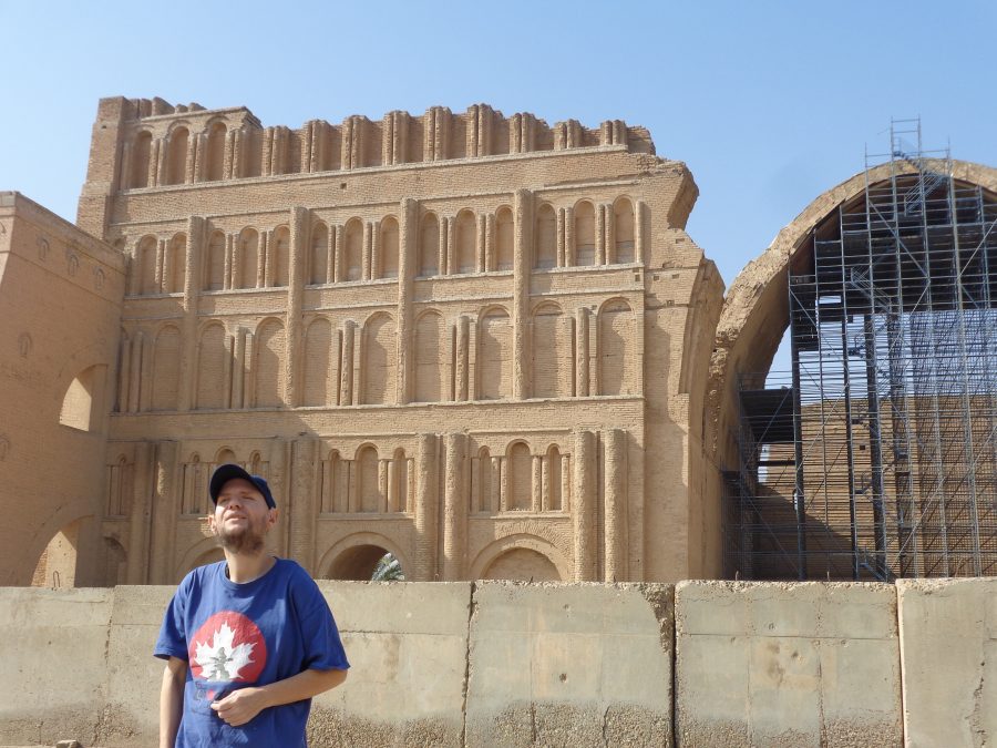 View showing part of the facade of the imperial palace at Taq Kasra with the huge brick arch to the right. To the left an arch passes through the thick brick walls at ground level. On the far left a large brick buttress extends out forwards from the corner of the towering wall: this was likely built to give the structure extra strength. Tony is again stood in the foreground with a concrete wall behind.