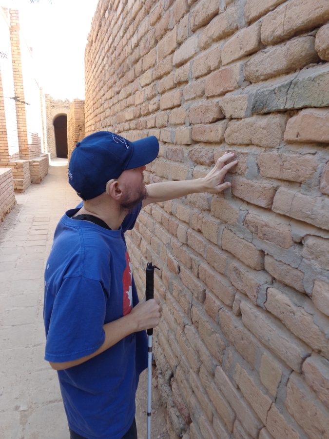Tony stood in a narrow passageway between high mud-brick walls. Tony is touching the rough texture of one of the tan-coloured brick walls. The bricks are quite uneven in size and shape. An arched doorway can be seen at the end of the passageway. This shot was taken in one of the four excavated and partially restored temples that are located close to the Ziggurat.