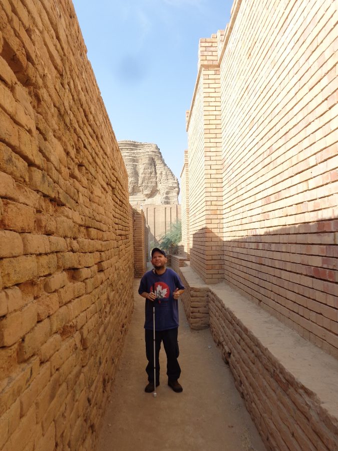 Tony standing in another passageway or alleyway within the complex of temples close to the Ziggurat of Dur-Kurigalzu. The Ziggurat itself can be seen towering above in the middle distance. The right wall is made-up of modern regularly-shaped mud bricks while the left wall is more irregular and weathered looking.