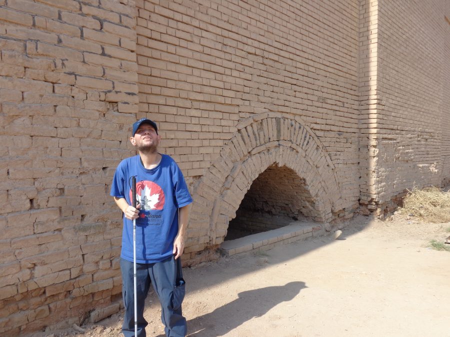 Tony stood in front of a tall light grey-brown brick wall. The bricks near the ground look old and could date from ancient Babylon, while those higher up look modern, and are likely from reconstruction. Next to Tony is a substantial arched opening in the wall at close to ground level. The purpose of the opening is unclear, it could be a tunnel, an entrance to an underground chamber, or a now partially submerged gate into the ancient city.