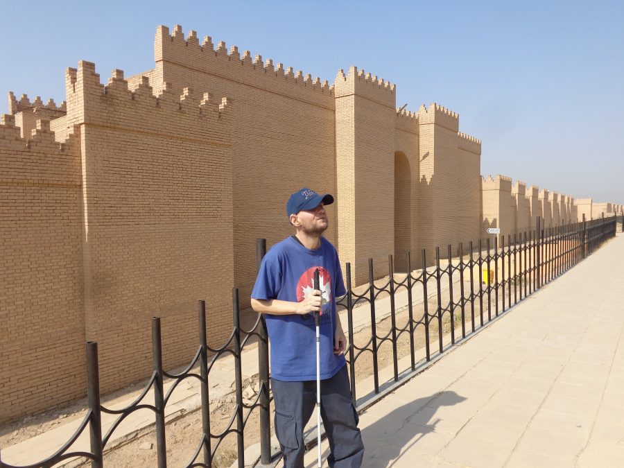 A tall tan-coloured brick defensive wall with rectangular towers and a crenellated top. The wall stretches into the distance, with a large arched gateway a little way in front. Tony is stood by black metal railings with a repeating curved pattern, which at this location runs parallel to the wall. The wall appears to be a modern reconstruction of the ancient city walls of Babylon.