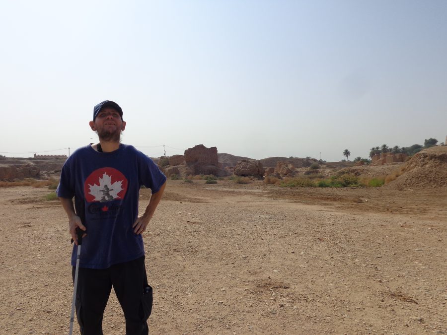 Tony stood in the ruins of ancient Babylon. In the background fragments of substantial mud-brick walls can be seen. There are also piles of rubble probably where walls have collapsed or been destroyed. The foreground is mostly flat with the ground comprised of gravel and sand. Palm trees can be seen in the middle distance. The site of ancient Babylon avoided occupation and damage by ISIS but was previously damaged by both the US army and militants following the 2003 invasion of Iraq. The US forces built a military base in the middle of the ancient site and in the process crushed archaeological remains.