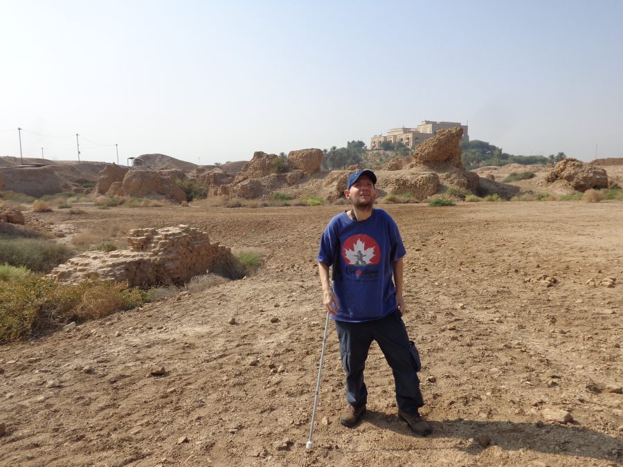 Tony stood in the dry, rocky landscape of ancient Babylon. Several fragments of crumbling mud-brick walls can be seen. Some of these walls appear to be a metre or more in thickness. Tony is in the foreground wearing a blue cap and tee-shirt and holding his white mobility cane. Beyond the ruins, a large modern building can be seen, on a man-made hill overlooking the site. This is a palace built by Saddam Hussein in the 1980s. The former Iraqi president built dozens of palaces during his 24 years as head of state. There are plans to turn the empty palace into an archaeological museum.