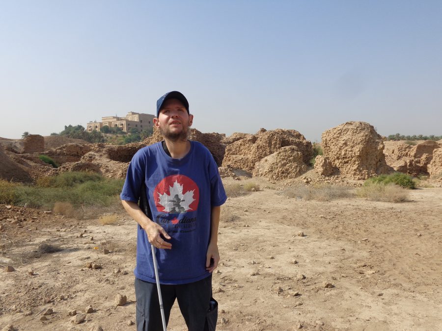  Another shot of Tony amongst the ruins of ancient Babylon. Dry scrubby vegetation can be seen growing around the crumbling mud-brick walls. Saddam Hussein's palace can again be seen on the hill at the edge of the site. The palace's wall are tan coloured matching the colour of the mud bricks. Palms trees and other green vegetation can be seen growing on the palace hill. The palace appears to be mostly two or three storeys in height with large arched windows.