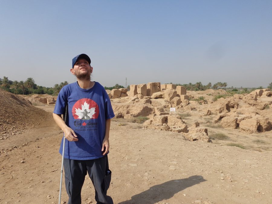 More ancient mud brick walls spread out behind Tony who is stood in the foreground. The tallest fragments of wall appear to be two or three metres in height. The landscape here is uneven with many mounds and hollows. Further in the distance there is a cluster of green palm trees contrasting with the arid grey-brown foreground.