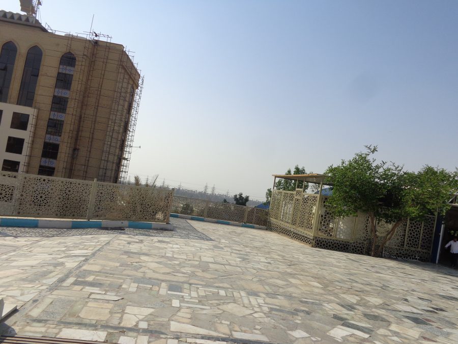 A raised courtyard located close to the Imam Ali Shrine. The surface is paved with irregular stone tiles. Metal railings with geometric decoration forming star patterns line the sides of the courtyard. Trees and pylons can be seen away in the distance. There is a pavilion-like structure to the right, and off to the left, a tall building is under construction.