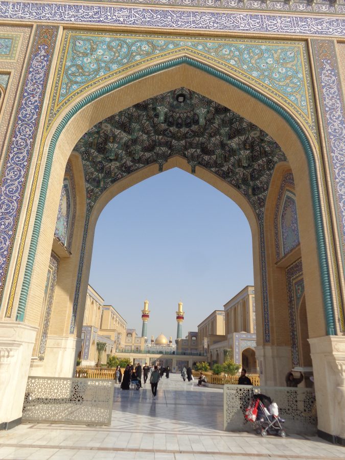 View through a large monumental gateway leading into the extended complex of buildings containing the Imam Ali Shrine. The gateway is covered with intricate tilework predominantly in blue, gold and turquoise colours and including stylised designs depicting stems, leaves and flowers. There are also bands of calligraphy in Arabic script. The gateway is formed of a very tall pointed arch with the interior also elaborately decorated with tiles. Beyond the archway there is a long walkway which passes through a series of courtyards before reaching the Imam Ali Shrine and Mosque itself at the far end of the complex. The closest courtyard is paved with marble and contains plants and fountains. A pair of tall gold-topped minarets can be seen mid-way through the complex. In the distance the large gold dome belonging to the Imam Ali Shrine can be seen as well as another pair of minarets also attached to the shrine itself.

The Imam Ali Shrine is a monumental Islamic mausoleum and mosque located in Najaf, Iraq. It is the burial site of Ali ibn Abi Talib who was the cousin and son-in-law of the Prophet Muhammad and the first Imam of Shia Islam.  Shia Muslims consider the site to be the fourth holiest in Islam and on average it is visited by over 8 million pilgrims annually. The first structure was built over the tomb in 977 CE by the Buyid dynasty. It has been extensively expanded and restored by various empires throughout the centuries, including the Safavids and the Ottomans.