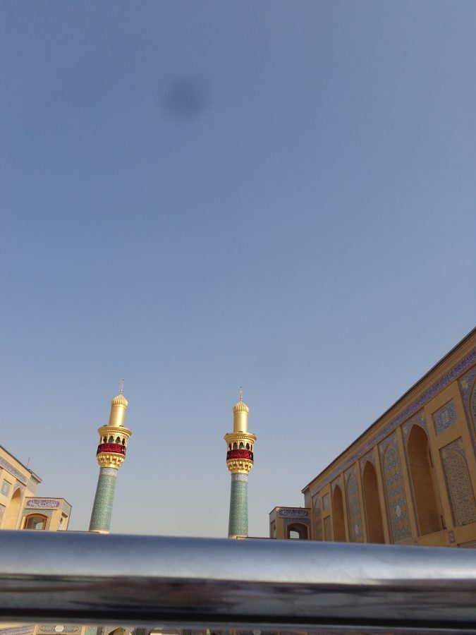 View over metal railings within the complex containing the Imam Ali Shrine. In front a pair of tall minarets can be seen. These minarets are additional to the pair that belong to the shrine itself. The top of these minarets are gold, including the balcony where a muezzin (a chosen person at a mosque would traditionally performs the adhan : the Islamic call to prayer). The balcony also has a band of crimson fabric wrapped around it with includes Islamic calligraphy. Lower down the minarets are covered with turquoise tilework with geometric decoration and another band of calligraphy at the top. The buildings at the sides are built in traditional style with tall arched window recesses, blocks of geometric tilework and bands of calligraphy along the top.