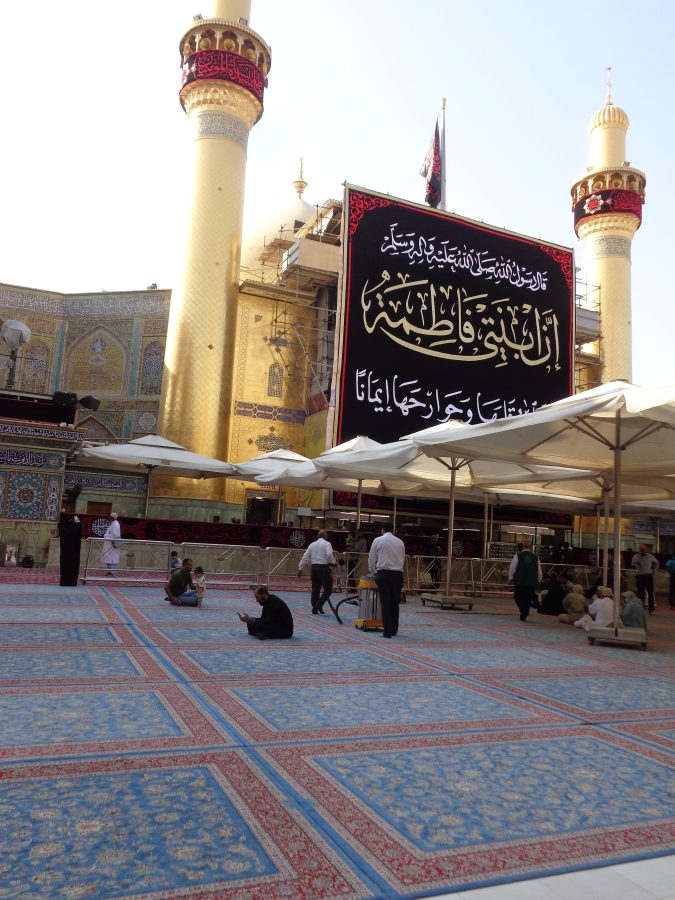 Outside the Imam Ali Shrine and Mosque. The courtyard in front is covered with large blue and red traditional rugs and people can be seen sitting, perhaps praying or in contemplation.  This area is partially shaded with large sun umbrellas. The front facade of the mosque is clad almost entirely in gold including the pair of minarets at the sides. A large front section of the mosque is temporarily obscured by scaffolding and this is covered with a large banner with writing in Arabic script on a black background. Only a small part of the mosque's large gold dome is visible at roof level.