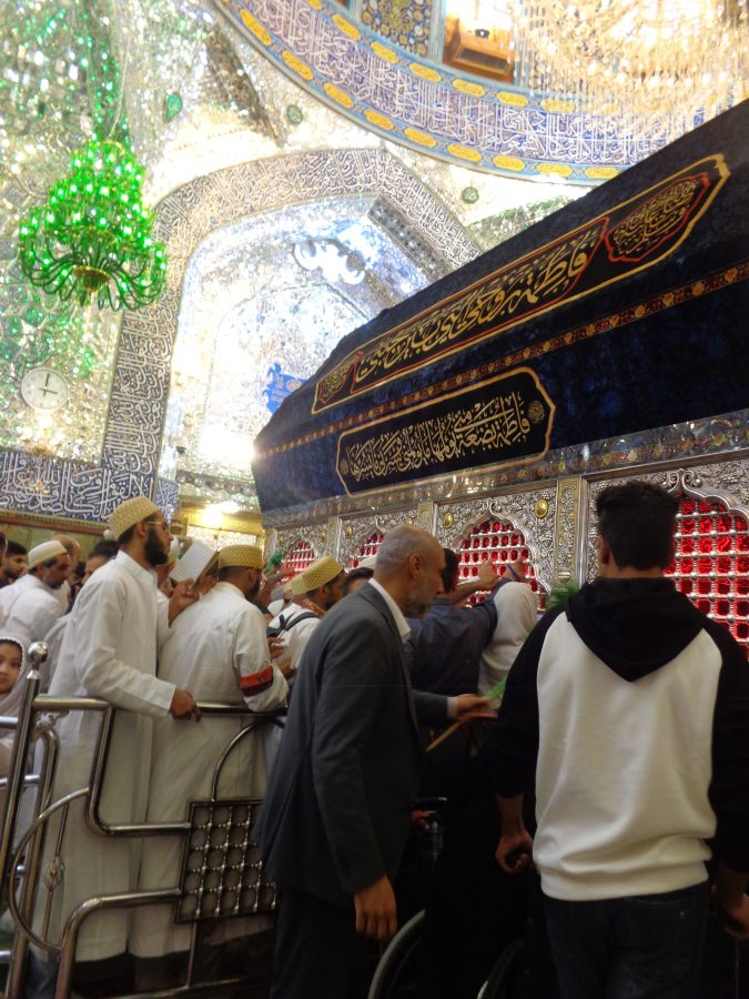 Inside the Imam Ali Shrine with the tomb of Ali ibn Abi Talib (also known as Imam Ali) in front. A group of people, predominantly men, many in white robes and with cream flat-topped kufi hats, are paying their respects in front. The outer structure of the tomb is ornately decorated in silver and gold with stylised decoration including leaves, stems and berries. The top of the tomb is covered with a deep blue fabric with bands of calligraphy on it. The surrounding structure of the shrine is also very richly decorated. There are several large chandeliers including a green in front. The walls have wide bands of calligraphy in blue and white and other elaborate decoration that reflects the light.