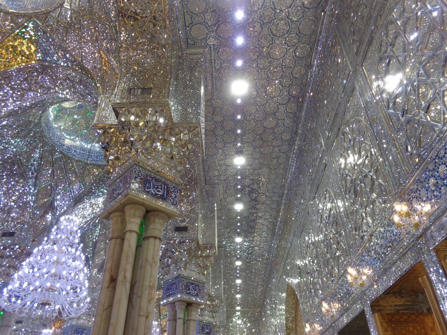 View of the glistening decoration inside the Imam Ali Shrine. There are marble columns and chandeliers. The walls and ceiling are completely covered with geometric decoration reflecting the light in silver, gold and blue. The decoration appears to contain large areas of inlaid crystal, perhaps glass or precious stones. The ceiling includes arches and domes all covered with the shimmering decoration.