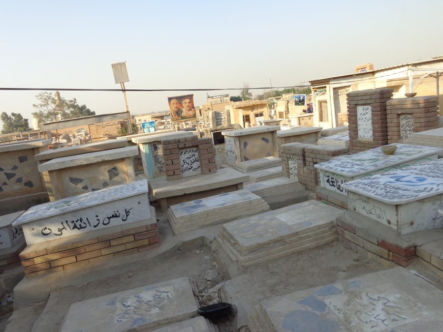 Looking across Wadi-us-Salaam cemetery with densely packed tombs extending out ahead. The size of the visible tombs varies from small structures close to the ground through to substantial flat roofed buildings that appear to be mausolea or crypts. A few of the tombs have attached banners, which display photos or portraits, presumably of the person buried there. Tall trees and green foliage can be seen in the distance.