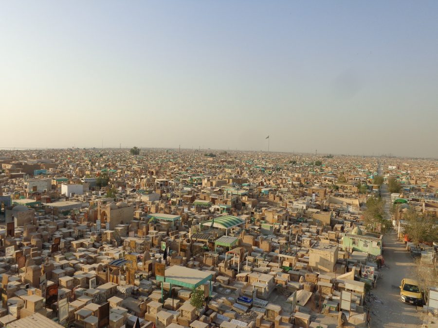 Again looking down over Wadi-us-Salaam cemetery. A long straight road extends through the cemetery into the distance. The structures in the cemetery are densely packed and vary from individual tombs through to large buildings containing crypts. In the distance, outside the cemetery, a very tall flag pole can be seen. A flag is flying at the top but it is too distant to identify. 