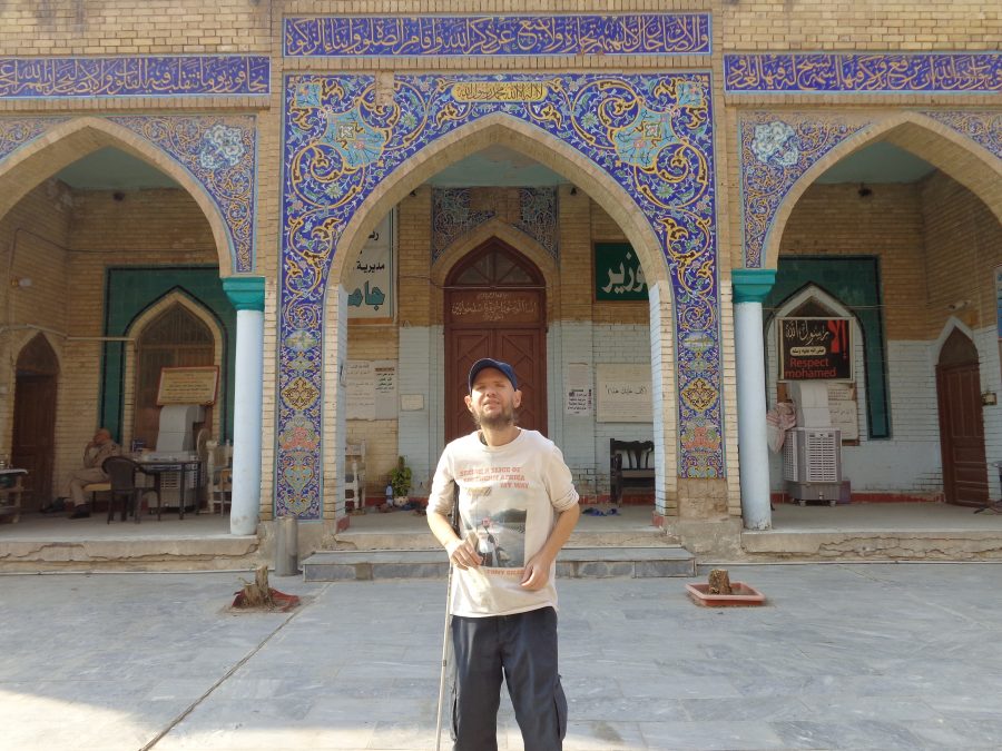 Tony outside the ornate entrance to the Al-Wazeer (Al-Wasir) Mosque which includes Islamic and Ottoman architectural features. Three pointed-arches are in view which lead into a large porch with the wooden main doorway in the centre. The outer facade is decorated with intricate tile work. At the top of the arches is blue and gold writing in Arabic script. Below this the tiles depict stylised plants with stems, leaves and flowers in several colours including blue, gold, green and turquoise. Within the porch there are various signs and posters: the only one in English reads "Respect Mohamed". A man, perhaps a guard or caretaker, can be seen sitting in a chair. This Sunni Islam mosque was built by the Wazir (governor) of Baghdad, Hassan Pasha, in 1599. It is situated behind the Souk al-Sarai near the riverbank of the Tigris. 