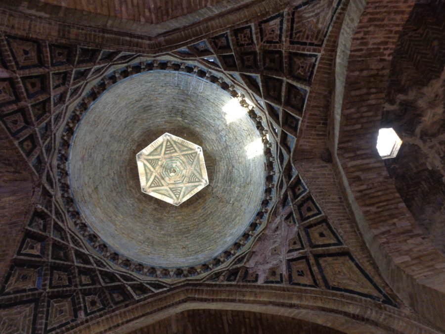The Al-Wazeer (Al-Wasir) Mosque's impressive brick domed ceiling with intricate geometric patterns. There is a large central dome that is supported by arches beneath. The top of the dome has a large hexagonal opening with light shining from hidden windows at the sides. Within the hexagonal opening there is a decorative brickwork design forming a large six-pointed star. Below the circular base of the dome the brickwork forms a pattern made-up of four-sided kite shapes: the size and angles of this shape change as the pattern extends down the sides of the arches. In addition, to the right, a hexagonal window can be seen in the vaulted brick ceiling. The architecture showcases classic Islamic or Middle Eastern design elements.
