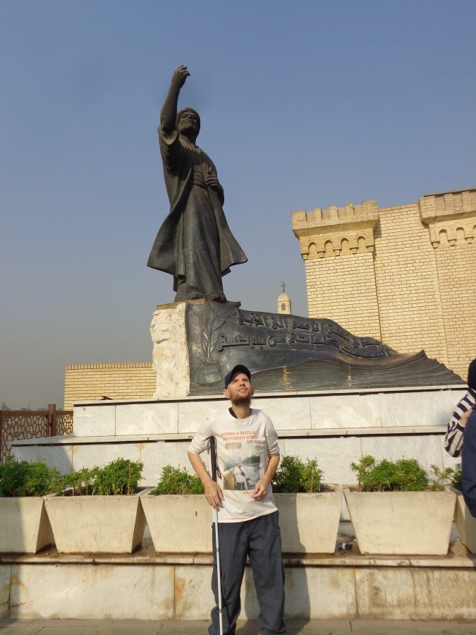 Tony stood in front of a large statue of Al-Mutanabbi. This bronze statue depicts a robed Mutanabbi with one arm raised upwards and the other at his chest. Al-Mutanabbi was born in circa 915 and died in An Numaniyah in 965 aged about 50. He was an Abbasid-era Arab poet at the court of the Hamdanid emir Sayf al-Dawla in Aleppo. In front of the statue are stone containers filled with plants with green foliage. In the background are beige brick buildings and the domed top of the Qushla Clock Tower can also be seen. 