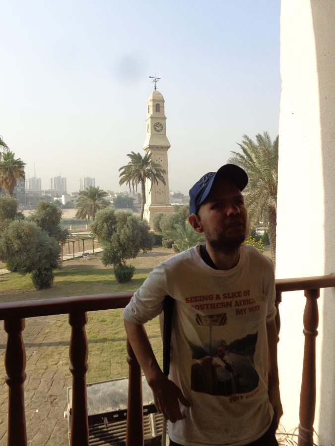 Tony on a balcony in the Qushla building. Behind him are wooden railings and below is a garden covered with grass as well as several palm and olive trees. Just beyond this stands the 19th-century Qushla Clock Tower and then the banks of the Tigris river. Construction of the Qushla (or Qishleh) began in 1881 under Ottoman governor Mehmed Namık Pasha. It was built as headquarters and barracks for the Ottoman army who were responsible for protecting and providing security for Baghdad at that time. The Qushla Clock Tower was completed in 1871 and was built to wake the soldiers housed within the Qushla. It is 30 metres in height. There are clock faces on all four sides of the tower. The current clock faces and clock mechanism were a gift from King George V to King Faisal I when he visited Britain in 1927.