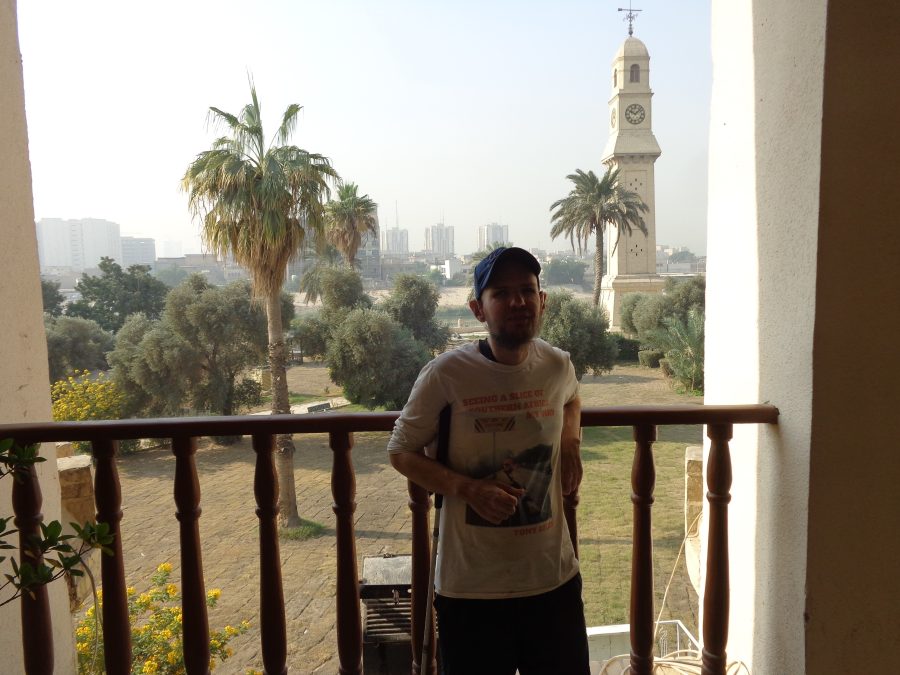Tony again on the balcony of the Qushla with the Qushla Clock Tower behind. The clock tower is topped with a weather vane. Several tall buildings are visible away on the far side of the Tigris river. 