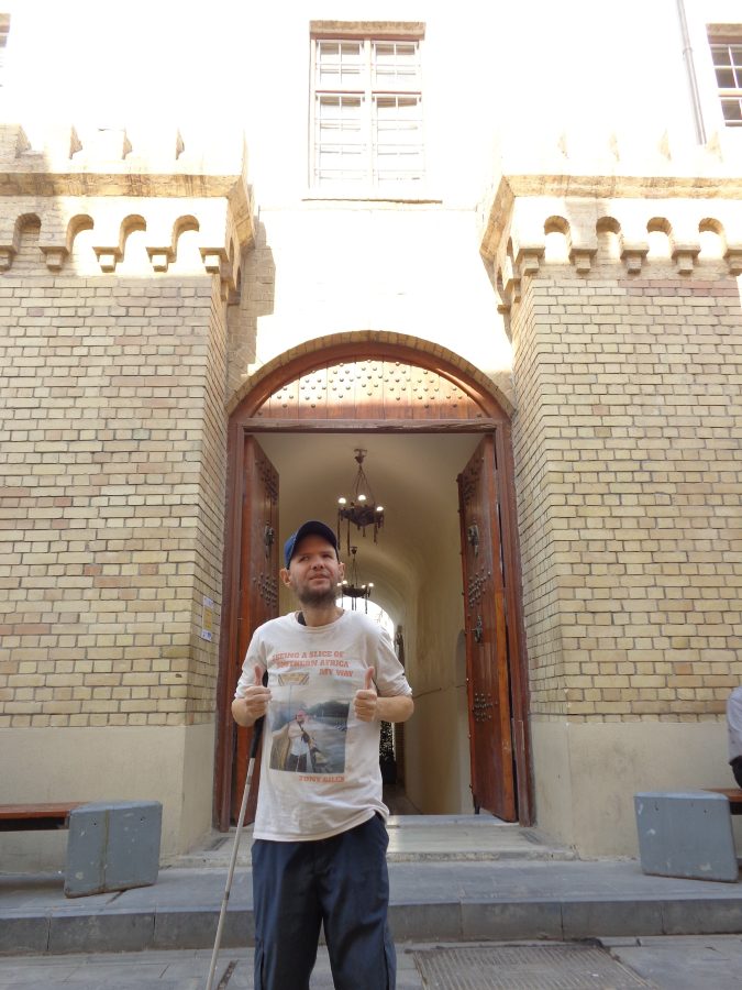 Tony stood in front of the arched entrance to the Qushla. A set of large double doors are open: they are made of thick wooden with metal studs and knockers. The surrounding walls are made of beige brick.