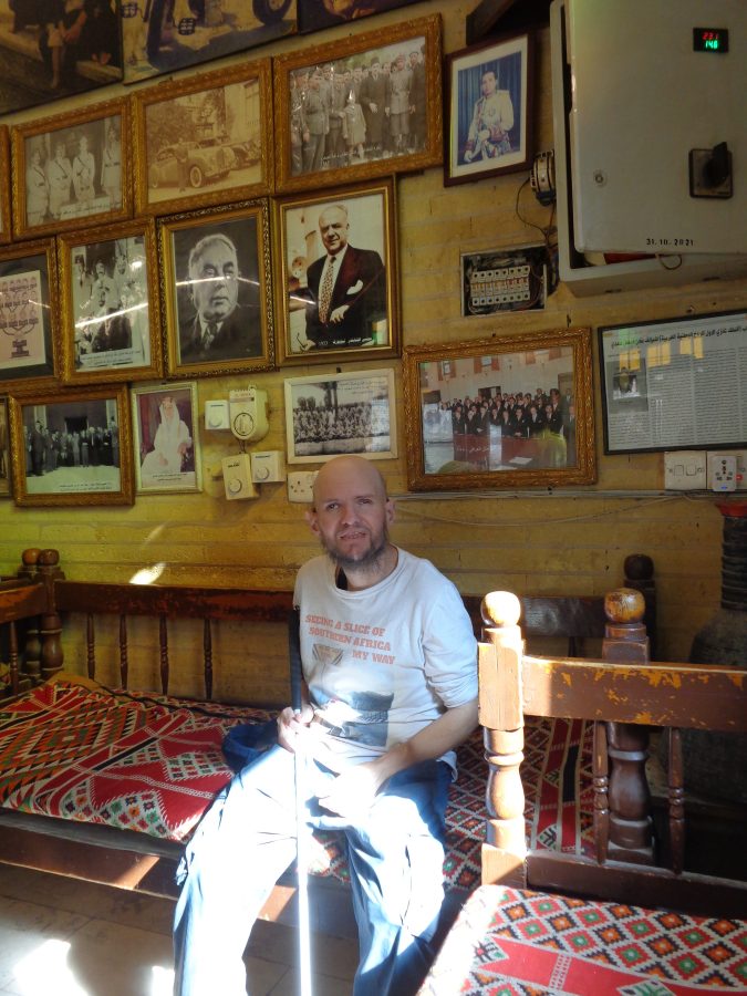 Tony sat on a traditional wooden bench covered with a colourful patterned fabric in the famous Shabandar Café. A yellow brick wall behind is filled with framed photographs, portraits, and documents, arranged in a collage-like manner. Most of the photos are black and white or sepia-toned and many of them show famous Iraqis who have visited this café, which is one of the oldest and most famous coffeehouses in Baghdad. During its history it has been frequented by poets, writers, politicians and intellectuals. It is located at the end of al-Mutanabbi Street near the Qushla. Originally a printing press, the coffee house was established in 1917. In the wake of sectarian violence following the US invasion of Iraq in 2003, the Shabandar Café was the victim of a car bomb attack in 2007, which killed more than a hundred people. The cafe was destroyed and four of owner Muhammad al-Khashali's sons were killed. The cafe subsequently rebuilt with the aim of preserving its cultural heritage and old identity with funding from the state, merchants, and its customers.