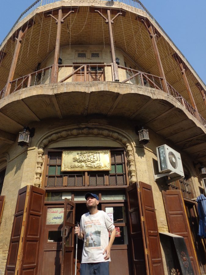 Tony outside the entrance of the Shabandar Café, a historic two-story building with a balcony spanning the structure at first-floor level. 