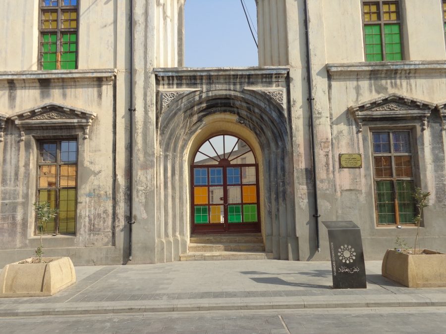 Outside the Old Baghdad Governorate Building. The building is in ruins with only the outer walls still standing. The front facade looks weathered but is mostly intact. There is a large arched doorway in the centre. The windows are now decorated with stained glass in green, yellow, and blue tones. The building was constructed in the first half of the 20th century and in 1934 it was renovated and rebuilt in English Victorian architectural style. Government offices were based here until 1985. It was damaged by fire and partially destroyed during the 2003 invasion of Iraq. The site is opposite the Qushla building off Mutanabbi Street.