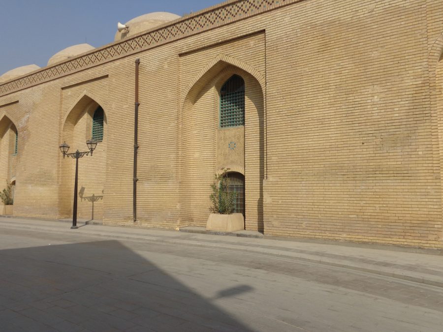 Yellow brick walls outside the Al-Sarai Mosque (King Ghazi Mosque). The mosque is historic but these outer walls look relatively modern. There are windows shaded inside large recesses topped with pointed arches. There are three small domes visible along the roof. Variously coloured bricks make a geometric pattern below the roof line. A traditional-style cast iron street lamp stands in the road in front.