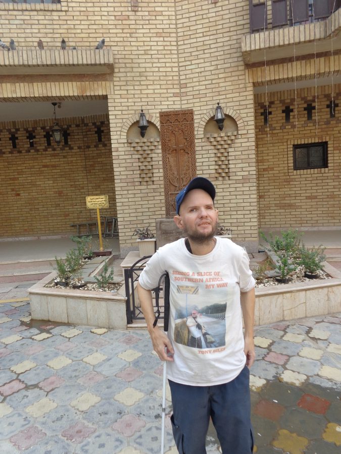 Tony stood in the courtyard of Sourp Asdvadzadzin (Holy Mother of God) Armenian church in Baghdad, which is also known as the Church of the Virgin Mary. It dates from 1639. In 1970 there was a renovation process and they discovered a box hidden in one of the walls that contained remains of 40 Armenian soldiers. The photo shows the brick walls of the church. There are some small raised flowerbeds in front as well as a tall rectangular stone carved with a geometric design around the edge, and in the middle, a cross surrounded by writing in Armenian script.
