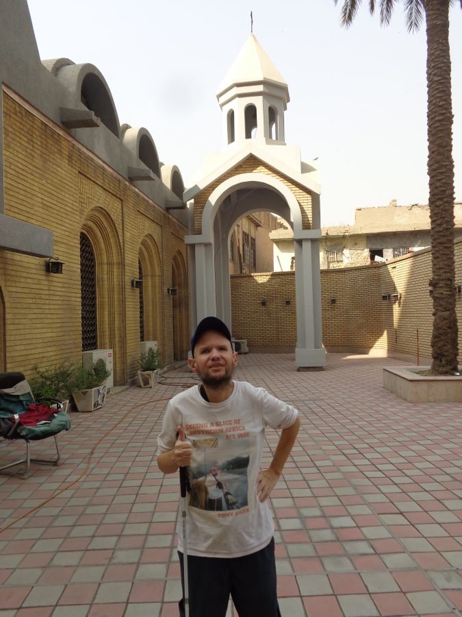 Tony again in the courtyard of Sourp Asdvadzadzin Armenian church. The brick outside walls of the church can be seen as well as a small free standing bell tower. The bell tower has a square profile with arched open sides that support a steeple and dome containing a bell. The bell is rung using a rope from ground level. The trunk of a tall palm tree is also in view. The courtyard is paved with pink and cream square blocks.