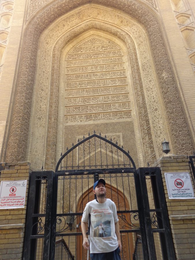 Tony stood in front of a grand doorway perhaps belonging to a palace. Above the wooden door itself is a very high pointed arch. The inside of the arch is covered with Arabic calligraphy and the sides are decorated with elaborate geometric designs. Access is blocked by metal railings and a sign says the site is closed for maintenance.